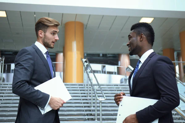 Dvě nadnárodní mladí podnikatelé mluví zároveň schody v moderní kancelářská budova. — Stock fotografie
