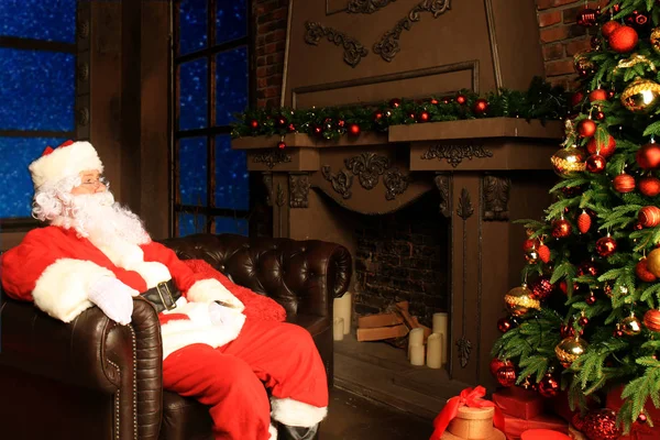 Papai Noel tendo um descanso em uma cadeira confortável perto da lareira em casa. — Fotografia de Stock