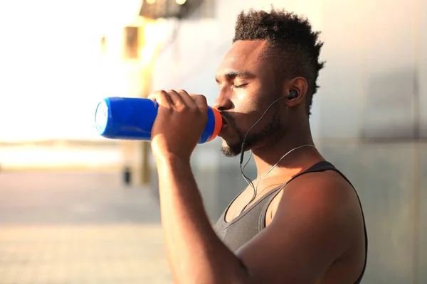 Hombre africano con ropa deportiva bebiendo agua mientras está parado afuera, al atardecer o al amanecer. Corredor . —  Fotos de Stock