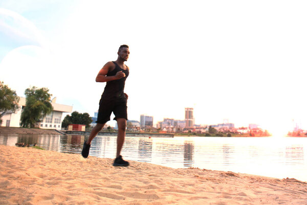 Full length of young African man in sports clothing jogging while exercising outdoors, in beach at sunset or sunrise