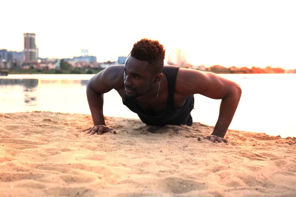 Hombre africano con ropa deportiva manteniendo la posición del tablón en la playa mientras hace ejercicio al aire libre, al atardecer o al amanecer . —  Fotos de Stock