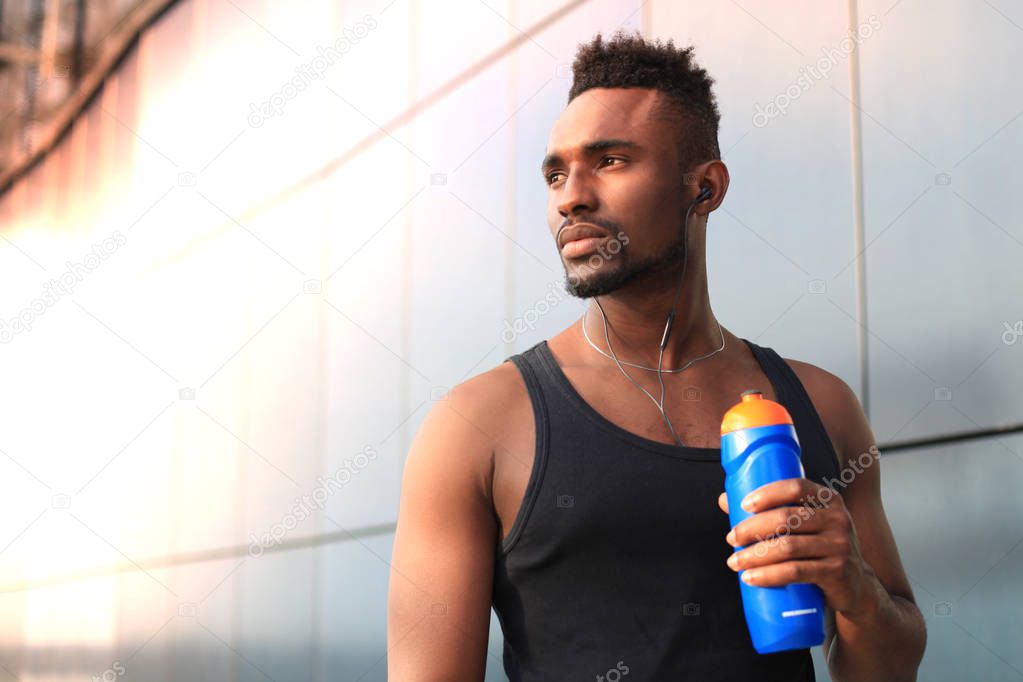 African man in sports clothing drinking water while standing outside, at sunset or sunrise. Runner.