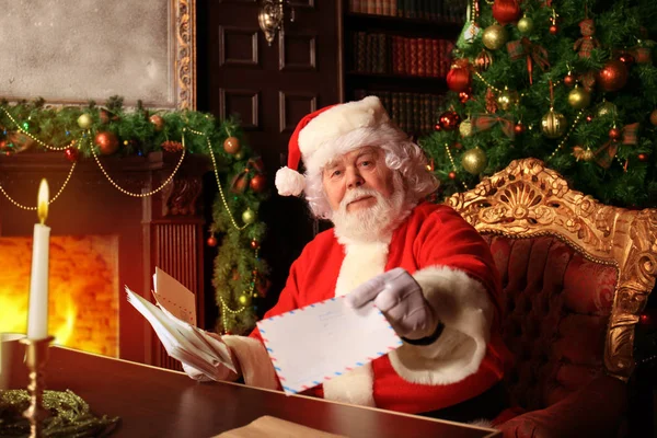 Mikołaj siedzi przy choince, trzyma świąteczne listy i odpoczywa przy kominku. — Zdjęcie stockowe