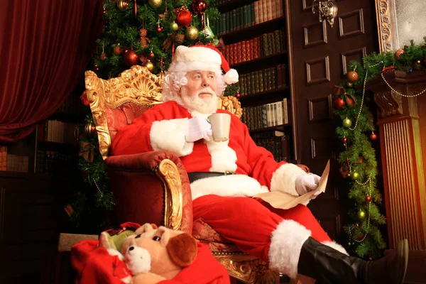 Portrait du Père Noël heureux assis dans sa chambre à la maison près de l'arbre de Noël et lisant la lettre de Noël ou la liste de souhaits. — Photo