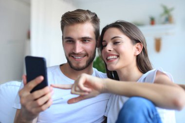 Genç çift evdeki kanepede oturmuş akıllı bir telefonla online içeriği izliyorlar..