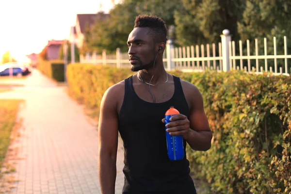 Hombre africano con ropa deportiva bebiendo agua mientras está parado afuera, al atardecer o al amanecer. Corredor . —  Fotos de Stock