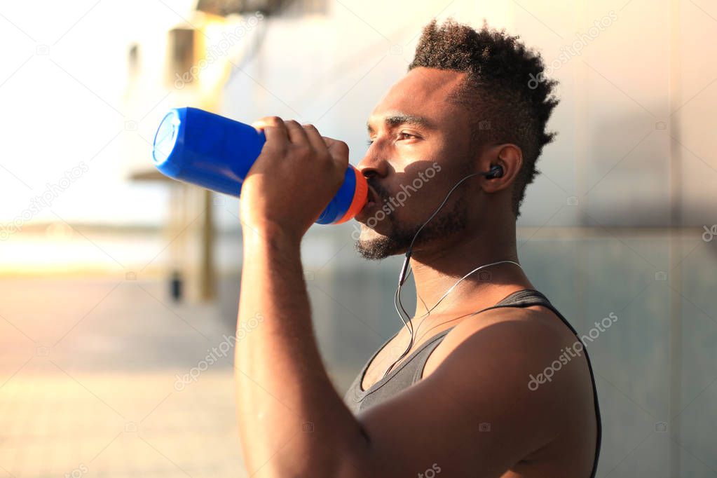 African man in sports clothing drinking water while standing outside, at sunset or sunrise. Runner.