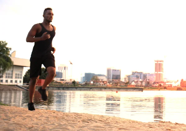 Longitud completa del joven africano en ropa deportiva trotando mientras hace ejercicio al aire libre, en la playa al atardecer o al amanecer . — Foto de Stock