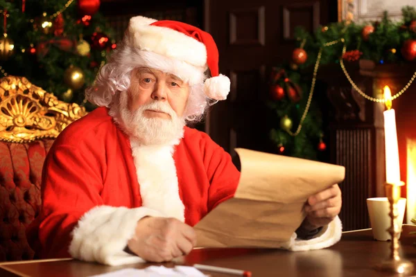 Retrato de Papai Noel feliz sentado em seu quarto em casa perto da árvore de Natal e lendo carta de Natal ou lista de desejos. — Fotografia de Stock