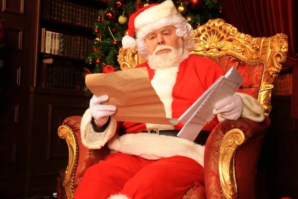 Retrato de Papá Noel feliz sentado en su habitación en casa cerca del árbol de Navidad y leyendo la carta de Navidad o la lista de deseos. — Foto de Stock