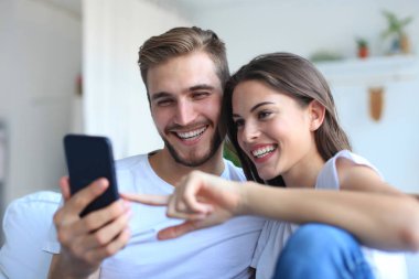 Genç çift evdeki kanepede oturmuş akıllı bir telefonla online içeriği izliyorlar..