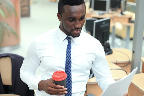 Succesvolle Afro-Amerikaanse zakenman lezen van papier op de werkplek, kijken naar financieel document, houden van koffie. — Stockfoto