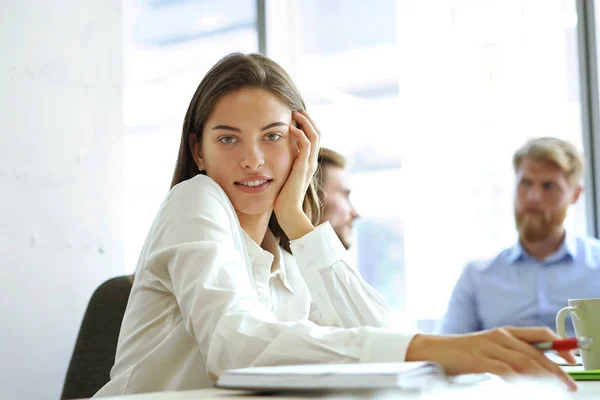 Designer i casual kläder sitter, tittar på kameran och leende, hans kolleger arbetar i bakgrunden. — Stockfoto