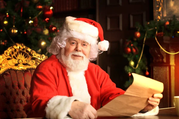 Retrato de Papai Noel feliz sentado em seu quarto em casa perto da árvore de Natal e lendo carta de Natal ou lista de desejos. — Fotografia de Stock