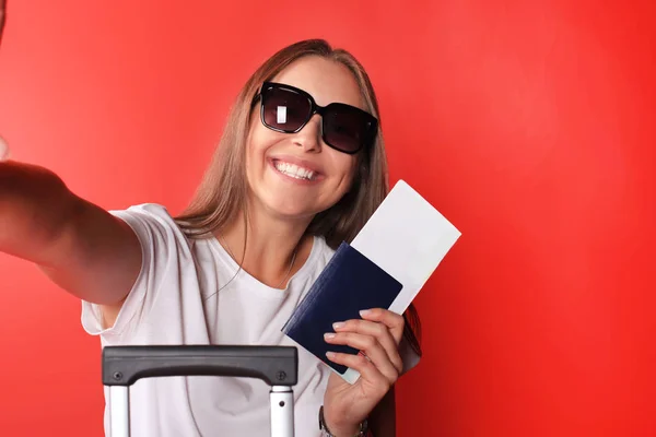 Femme souriante prenant un selfie avec des lunettes de soleil tout en tenant passeport avec valise rouge, isolé sur fond rouge . — Photo