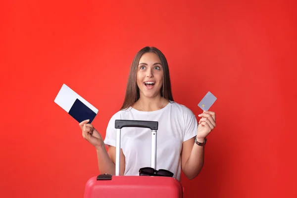 Fille touristique en été montrant carte de crédit en plastique, avec des lunettes de soleil, valise rouge, passeport isolé sur fond rouge . — Photo