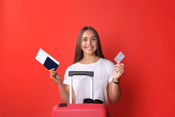 Fille touristique en été montrant carte de crédit en plastique, avec des lunettes de soleil, valise rouge, passeport isolé sur fond rouge . — Photo