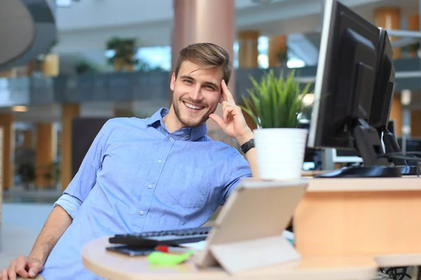 Portrait of happy man sitting at office desk, looking at camera, smiling.