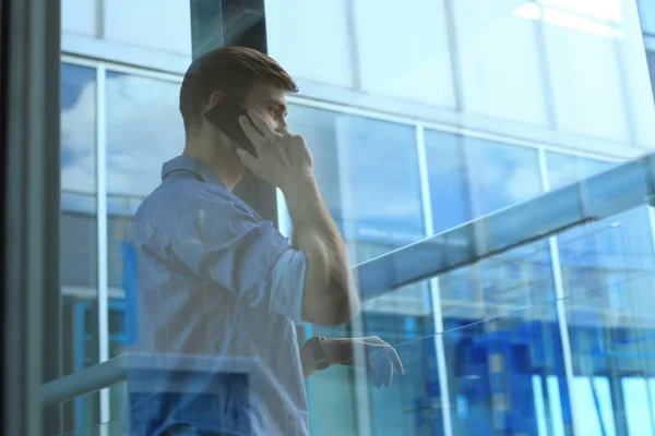 Empresário sorridente de pé e usando telefone celular no escritório . — Fotografia de Stock