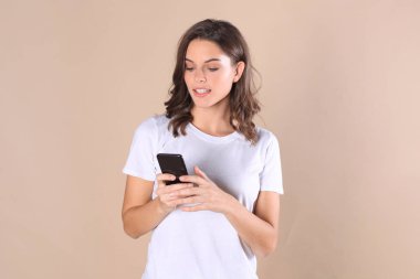 Cep telefonu kullaranarak temel giyim bej renkli arka plan üzerinde izole giymiş genç kadın.