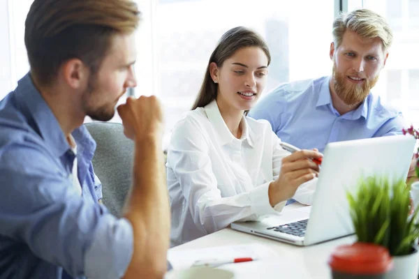 Groupe de jeunes gens modernes en tenue décontractée intelligente pointant vers un ordinateur portable et souriant tout en étant assis dans le bureau . — Photo