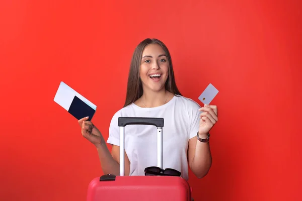 Fille touristique en été montrant carte de crédit en plastique, avec des lunettes de soleil, valise rouge, passeport isolé sur fond rouge . — Photo