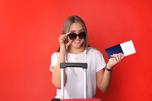 Menina turística jovem em roupas casuais de verão, com óculos de sol, mala vermelha, passaporte isolado no fundo vermelho . — Fotografia de Stock