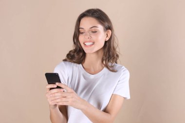 Cep telefonu kullaranarak temel giyim bej renkli arka plan üzerinde izole giymiş genç kadın.