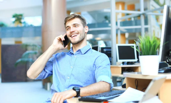 Hombre de negocios sonriente sentado y usando el teléfono móvil en la oficina . — Foto de Stock