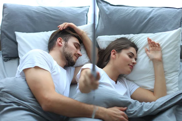 Beautiful young loving couple sleep and making selfie with stick in the bed.