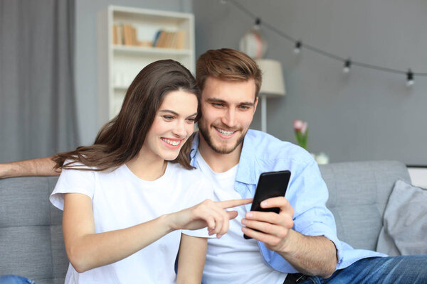 Young couple watching online content in a smart phone sitting on a sofa at home in the living room.