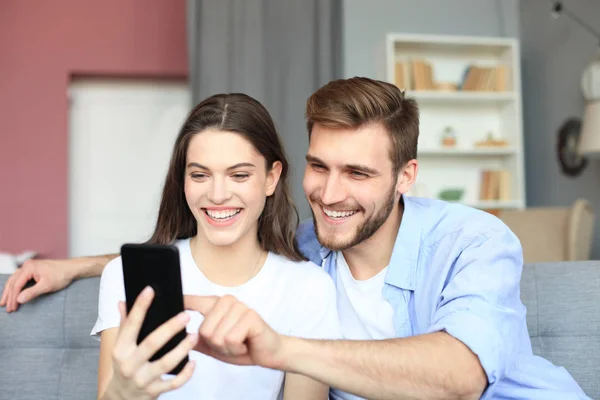 Pareja joven viendo contenido en línea en un teléfono inteligente sentado en un sofá en casa en la sala de estar. —  Fotos de Stock