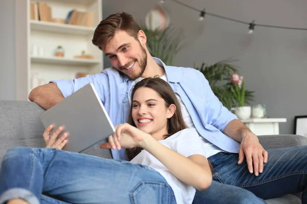 Pareja joven viendo contenido multimedia en línea en una tableta sentada en un sofá en la sala de estar. —  Fotos de Stock