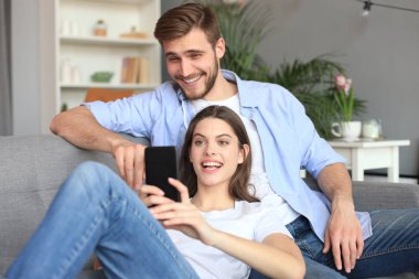 Genç çift evdeki kanepede oturmuş akıllı bir telefonla online içeriği izliyorlar..