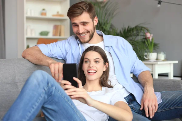 Pareja joven viendo contenido en línea en un teléfono inteligente sentado en un sofá en casa en la sala de estar. —  Fotos de Stock