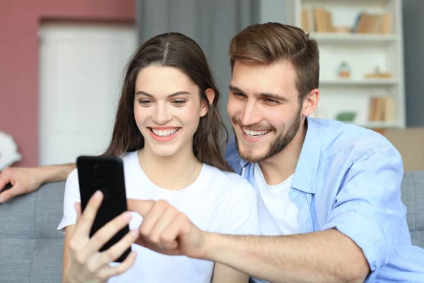 Giovane coppia che guarda contenuti online in uno smartphone seduto su un divano a casa in soggiorno. — Foto Stock