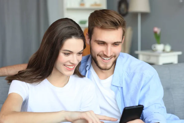 Giovane coppia che guarda contenuti online in uno smartphone seduto su un divano a casa in soggiorno. — Foto Stock