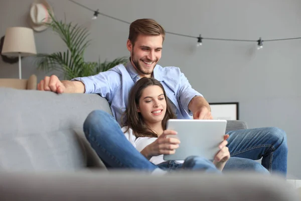 Pareja joven viendo contenido multimedia en línea en una tableta sentada en un sofá en la sala de estar. —  Fotos de Stock