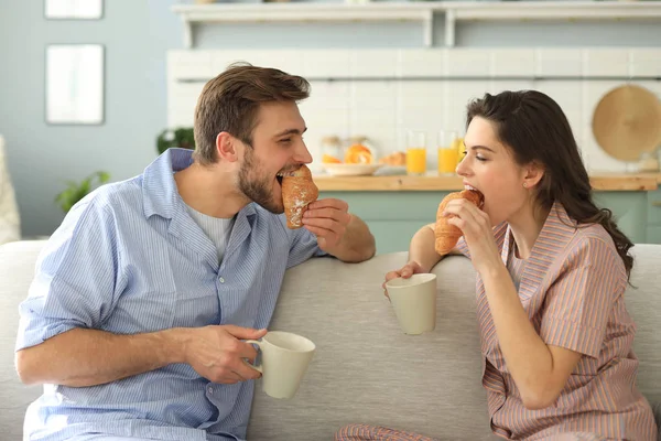 Happy young couple in pajamas in kitchen having breakfast, feeding each other a croissant. — Stock Photo, Image