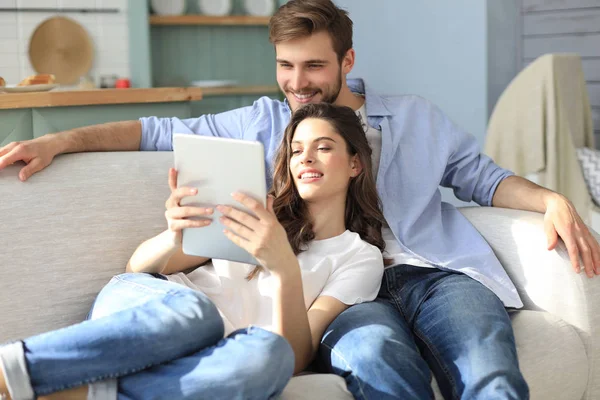 Pareja joven viendo contenido multimedia en línea en una tableta sentada en un sofá en la sala de estar. —  Fotos de Stock