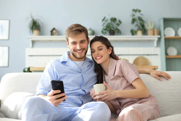 Pareja joven viendo contenido en línea en un teléfono inteligente sentado en un sofá en casa en la sala de estar. —  Fotos de Stock