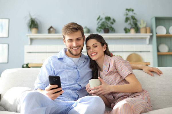 Young couple watching online content in a smart phone sitting on a sofa at home in the living room.