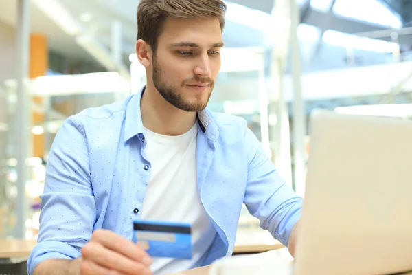 Hombre sonriente sentado en la oficina y paga con tarjeta de crédito con su computadora portátil. — Foto de Stock