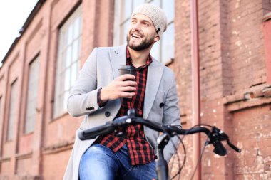 Gri ceketli ve şapkalı yakışıklı genç adam sokakta kahve içerken ayakta yürüyor.