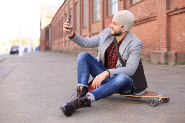 Gri ceketli ve şapkalı yakışıklı genç adam longboard'da oturuyor ve şehirde sokakta selfie çekiyor.
