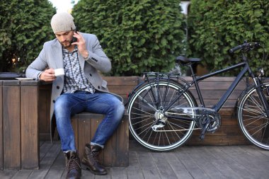 Gri ceketli ve şapkalı yakışıklı genç adam cep telefonuyla konuşuyor ve açık havada bisikletinin yanında otururken gülümsüyor.