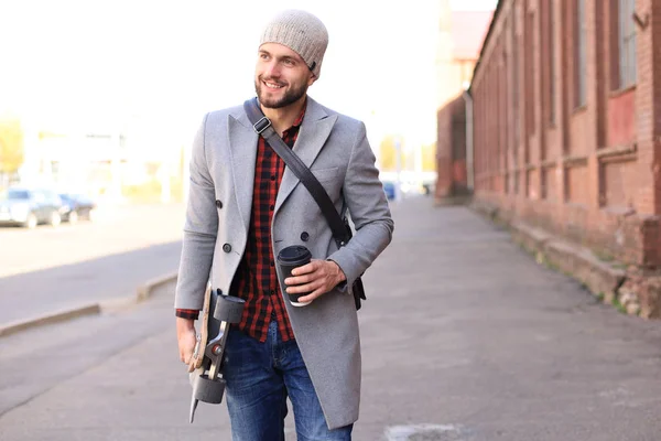 Joven guapo con abrigo gris y sombrero caminando por la calle, usando longboard . — Foto de Stock