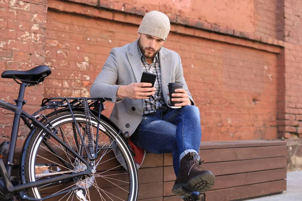Όμορφος νέος άντρας με γκρι παλτό και καπέλο που μιλάει στο κινητό τηλέφωνο και χαμογελάει ενώ κάθεται κοντά στο ποδήλατό του σε εξωτερικούς χώρους. — Φωτογραφία Αρχείου