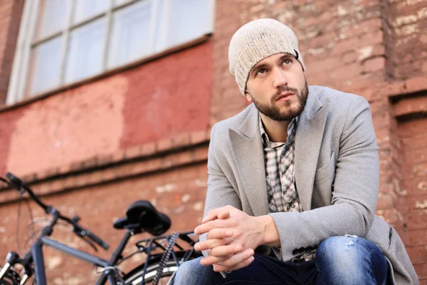 Bello giovane in cappotto grigio e cappello seduto su una panchina rilassato e pensare vicino alla sua bicicletta all'aperto . — Foto Stock