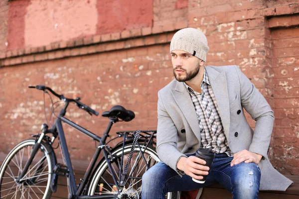 Beau jeune homme en manteau gris et chapeau assis sur un banc détendu en buvant du café et en pensant près de son vélo . — Photo